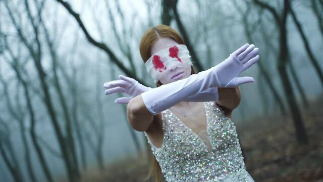 Scary zombie bride with bloody eyes under the bandage. Ghoul girl in a white dress in the forest in the fog in autumn