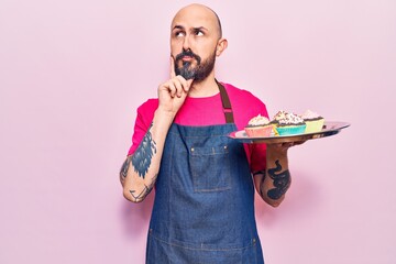 Young handsome man wearing apron holding cupcake serious face thinking about question with hand on...
