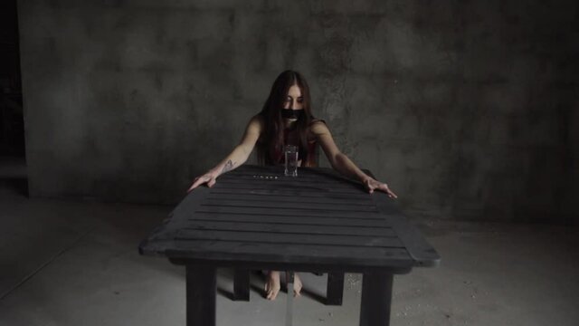 Young woman with gags in her mouth sits at table in dark room. There is a glass of water on the table.