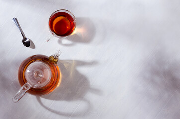 Cozy autumn atmosphere of morning teatime with black tea in transparent teapot with cup in orange sun beams and shadows on white wood table top view, copy space.