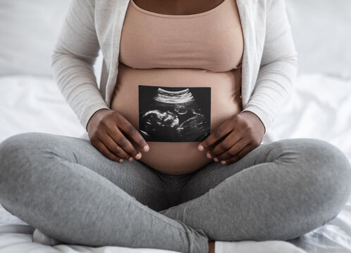 Unrecognizable black pregnant lady demonstrating her baby sonography photo, sitting on bed