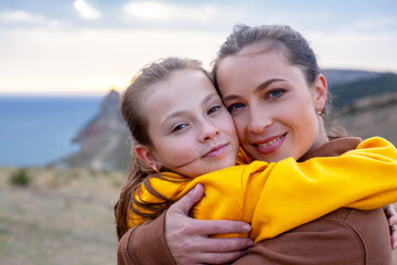 Mother and preteen daughter  wearing yellow and biege hoodie hugged on top of the mountain landscape and looking to the sea