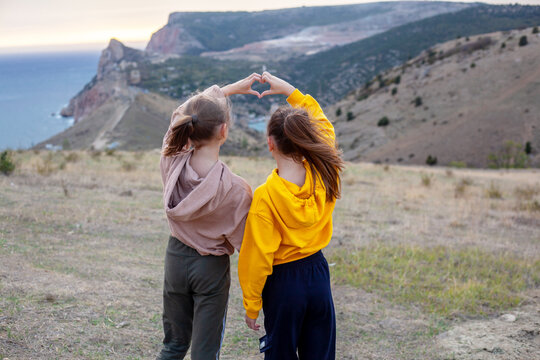 Two preteen girls traveler wearing yellow and biege hoodie walking  on top of the mountain landscape and looking to the sea. Kids making shape of love heart