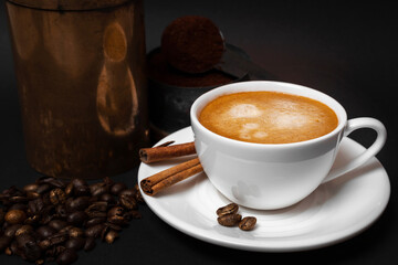 White cup of coffee, coffee beans, cinnamon sticks, aged coffee grinder, ground coffee in spoon. Close up