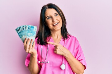 Middle age brunette doctor woman holding 100 brazilian reals banknotes smiling happy pointing with hand and finger