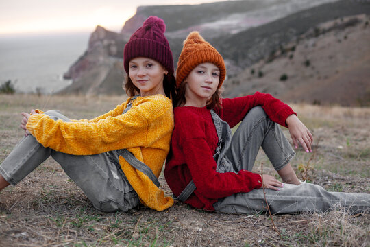 Two preteen girls traveler wearing denim overalls, yellow and red sweater and knitted hats sit on top of the mountain landscape and looking to the sea.