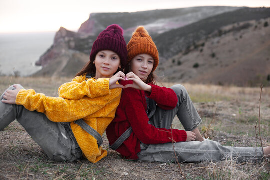 Two preteen girls traveler wearing denim overalls, yellow and red sweater and knitted hats sit on top of the mountain landscape and looking to the sea. Kids making shape of love heart