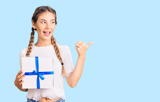 Beautiful caucasian woman with blonde hair holding gift pointing thumb up to the side smiling happy with open mouth