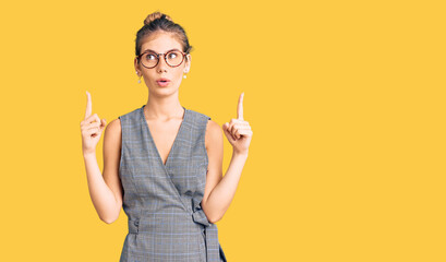 Beautiful caucasian woman with blonde hair wearing business clothes and glasses amazed and surprised looking up and pointing with fingers and raised arms.