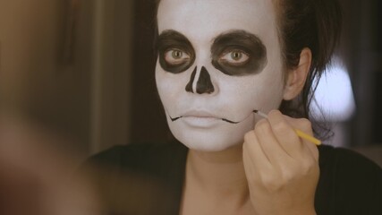 A brunette girl with a white tone on her face, a black nose and circles on her eyes with a thin brush draws arrows at the mouth. Close-up. Halloween makeup. October holiday.