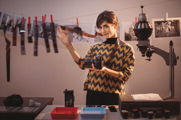 Happy young female holding filmstrip and smiling. Portrait of creative girl photographer in photo studio darkroom. Developing analog camera film