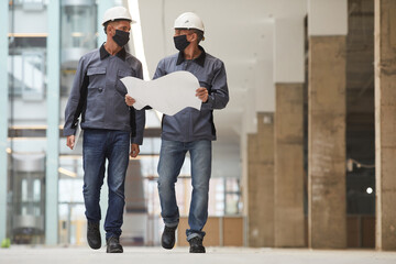 Full length portrait of two workers wearing masks and holding plans while walking towards camera at construction site, copy space