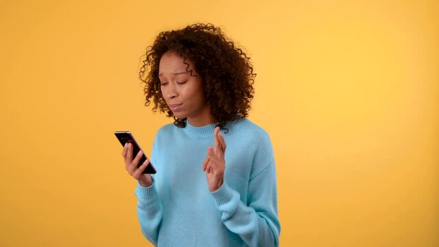 Good luck and winnings. young beautiful dark-skinned woman looks at the smartphone with great hope crossing her fingers. woman jumping for joy. in a blue sweater. person on isolated yellow background