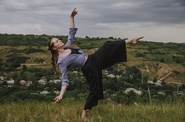Woman wearing large pants and striped shirt dancing with one leg up and hands in the air on a hill in a rural setting. Concept: self expression, freedom of movement, dance