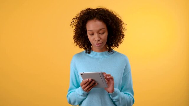 charming african american woman looks at the tablet. in a blue sweater. person on isolated yellow background. 4K