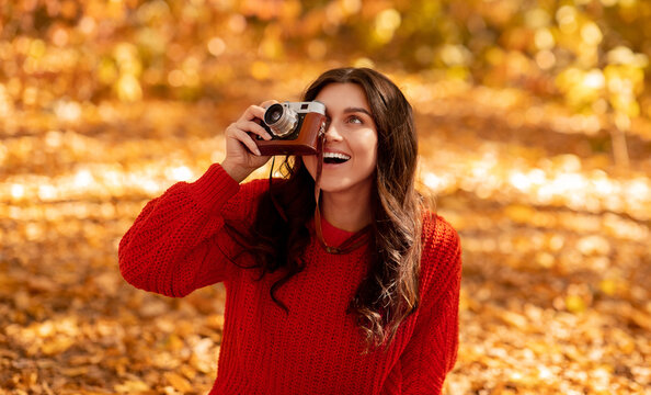 Smiling young woman in bright red sweater making pictures with retro camera at park on sunny fall day