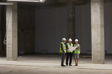 Wide angle portrait of business people wearing hardhats and holding plans while standing at construction site indoors, copy space