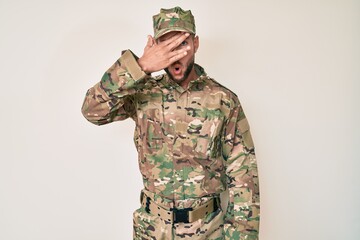 Young caucasian man wearing camouflage army uniform peeking in shock covering face and eyes with hand, looking through fingers with embarrassed expression.