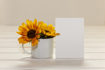 Blank white greeting card and enamel cup with sunflowers. Mockup on white wood table.