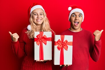 Young interracial couple wearing christmas hat and holding a gift pointing thumb up to the side smiling happy with open mouth
