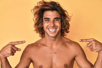 Young hispanic man standing shirtless looking confident with smile on face, pointing oneself with...