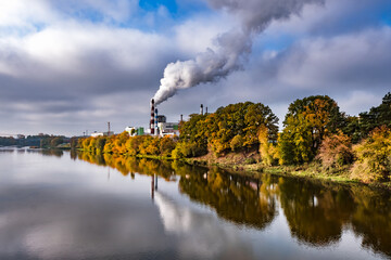 pipes of woodworking enterprise plant sawmill near river with autumn red yellow trees. Air pollution concept. Industrial landscape environmental pollution waste of thermal power plant