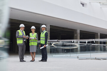 Full length portrait of three successful business people wearing hardhats and discussing investment deal while standing at construction site indoors, copy space