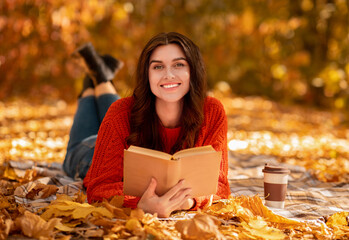 Smiling young woman with book and coffee to go lying on blanket among fallen autumn leaves and...