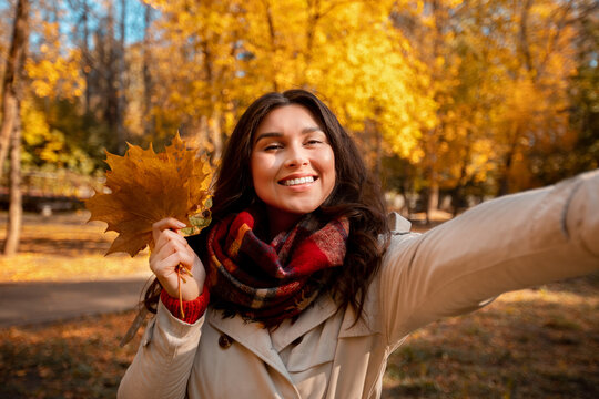 Beautiful young woman in fashionable fall clothes holding bouquet of autumn leaves and taking selfie at park