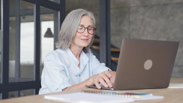 Smiling professional mature business woman wears glasses using laptop computer sits at workplace desk. Happy senior older employee 60s businesswoman executive working typing on pc at home from office.