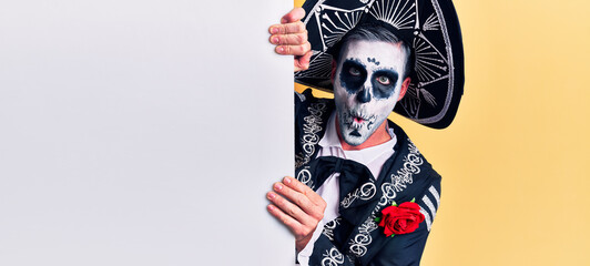 Young man wearing mexican day of the dead costume holding blank empty banner scared and amazed with open mouth for surprise, disbelief face