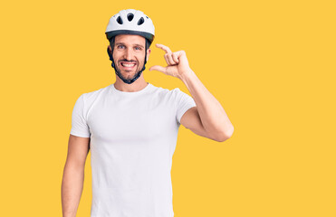 Young handsome man wearing bike helmet smiling and confident gesturing with hand doing small size sign with fingers looking and the camera. measure concept.