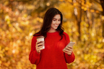 Lovely young lady in in warm knitted sweater using her smartphone and drinking takeout coffee at autumn park