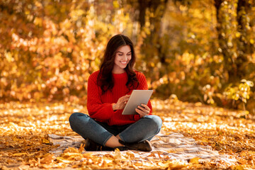 Pretty millennial woman in warm autumn outfit browsing internet on tablet computer at park