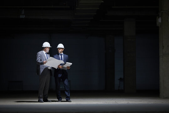 Dramatic Full Length Portrait Of Two Mature Business People Wearing Hardhats And Holding Plans While Standing In Dark At Construction Site Lit By Harsh Lighting, Copy Space