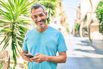 Middle age grey-haired man smiling happy using smartphone walking at street of city.
