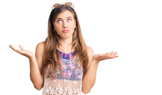 Beautiful caucasian young woman wearing bikini and sunglasses clueless and confused expression with arms and hands raised. doubt concept.