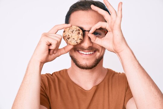 Young handsome man holding cookie smiling happy doing ok sign with hand on eye looking through fingers
