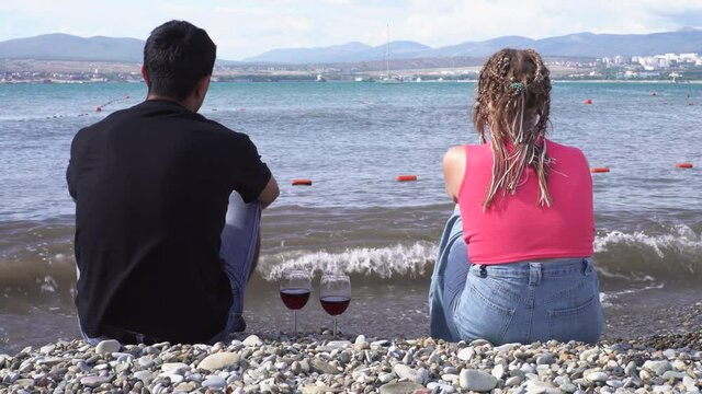 Rear view of a couple sitting at seaside with red wine glasses standing near on a summer sunny day. Media. Man in black shirt and woman with afro braids enjoying marine landscape.