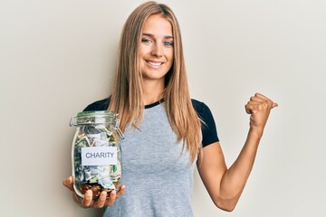 Obraz premium Young blonde woman holding charity jar with money pointing thumb up to the side smiling happy with open mouth