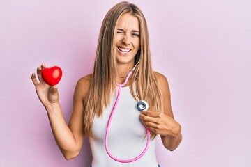 Young blonde woman using stethoscope holding heart winking looking at the camera with sexy...