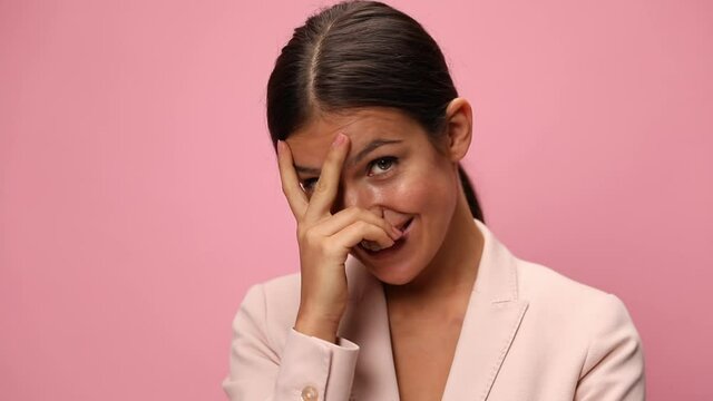 shy young businesswoman wearing pink suit, holding fingers on face, flirting and looking to side on pink background