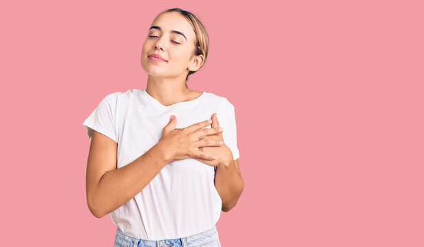 Young beautiful blonde woman wearing casual white tshirt smiling with hands on chest with closed eyes and grateful gesture on face. health concept.