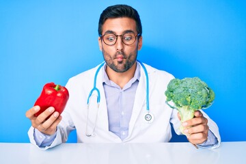 Young hispanic man wearing doctor uniform holding red pepper and broccoli making fish face with mouth and squinting eyes, crazy and comical.