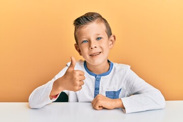 Adorable caucasian kid wearing casual clothes sitting on the table doing happy thumbs up gesture with hand. approving expression looking at the camera showing success.