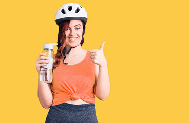 Young beautiful woman wearing bike helmet and holding water bottle smiling happy and positive, thumb up doing excellent and approval sign
