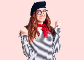 Young beautiful woman wearing french look with beret screaming proud, celebrating victory and success very excited with raised arms