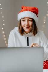 Young happy teen girl with excited face expression in red santa hat shopping online buying Christmas gifts paying by credit card using laptop seeing big sale discount.