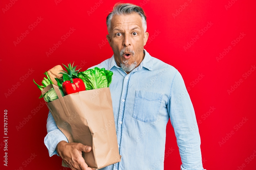 Sticker middle age grey-haired man holding paper bag with bread and groceries scared and amazed with open mo