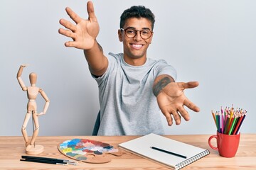 Young handsome african american man painter sitting palette and art manikin looking at the camera smiling with open arms for hug. cheerful expression embracing happiness.